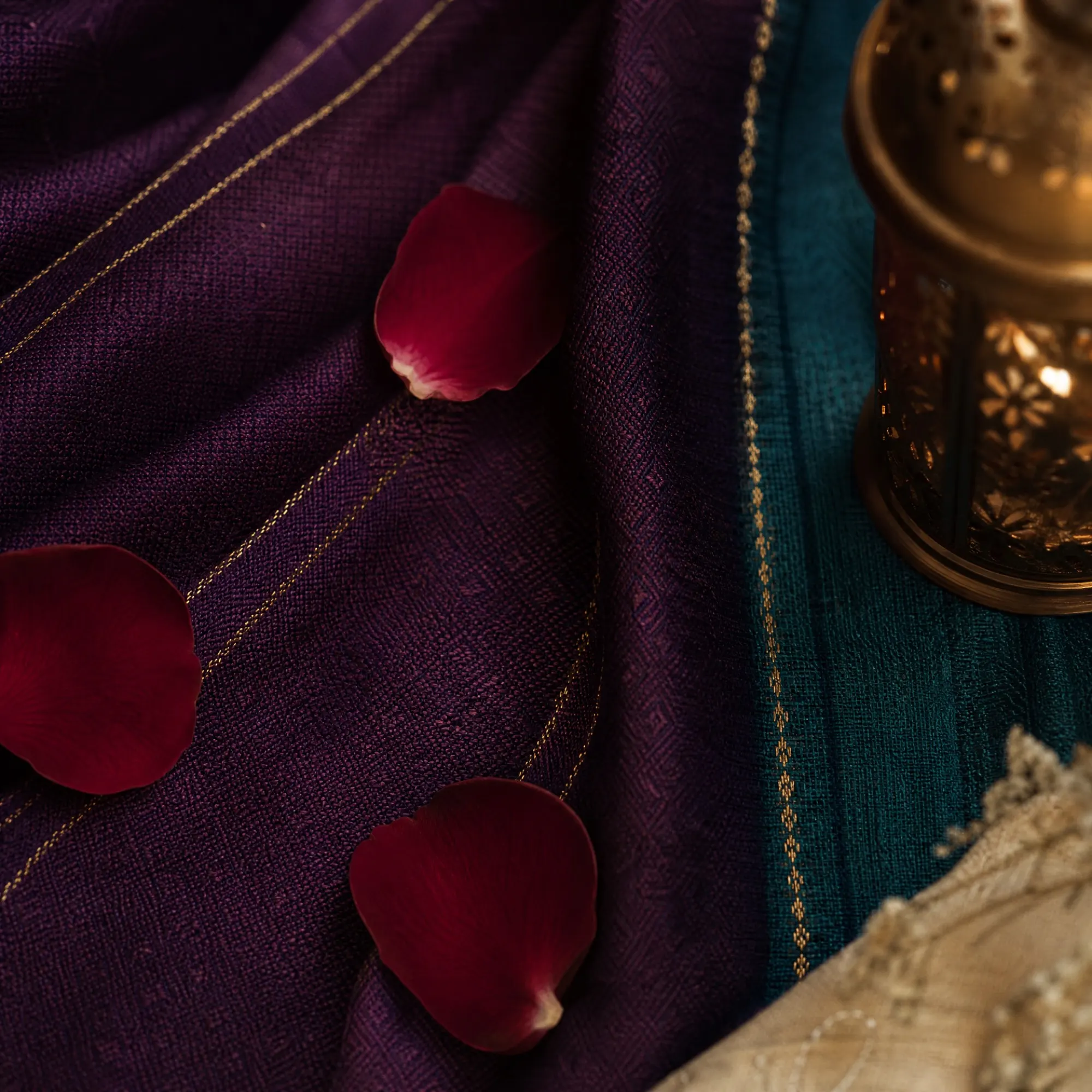 Petals drift across layered plum and teal cloth edged in gold thread.