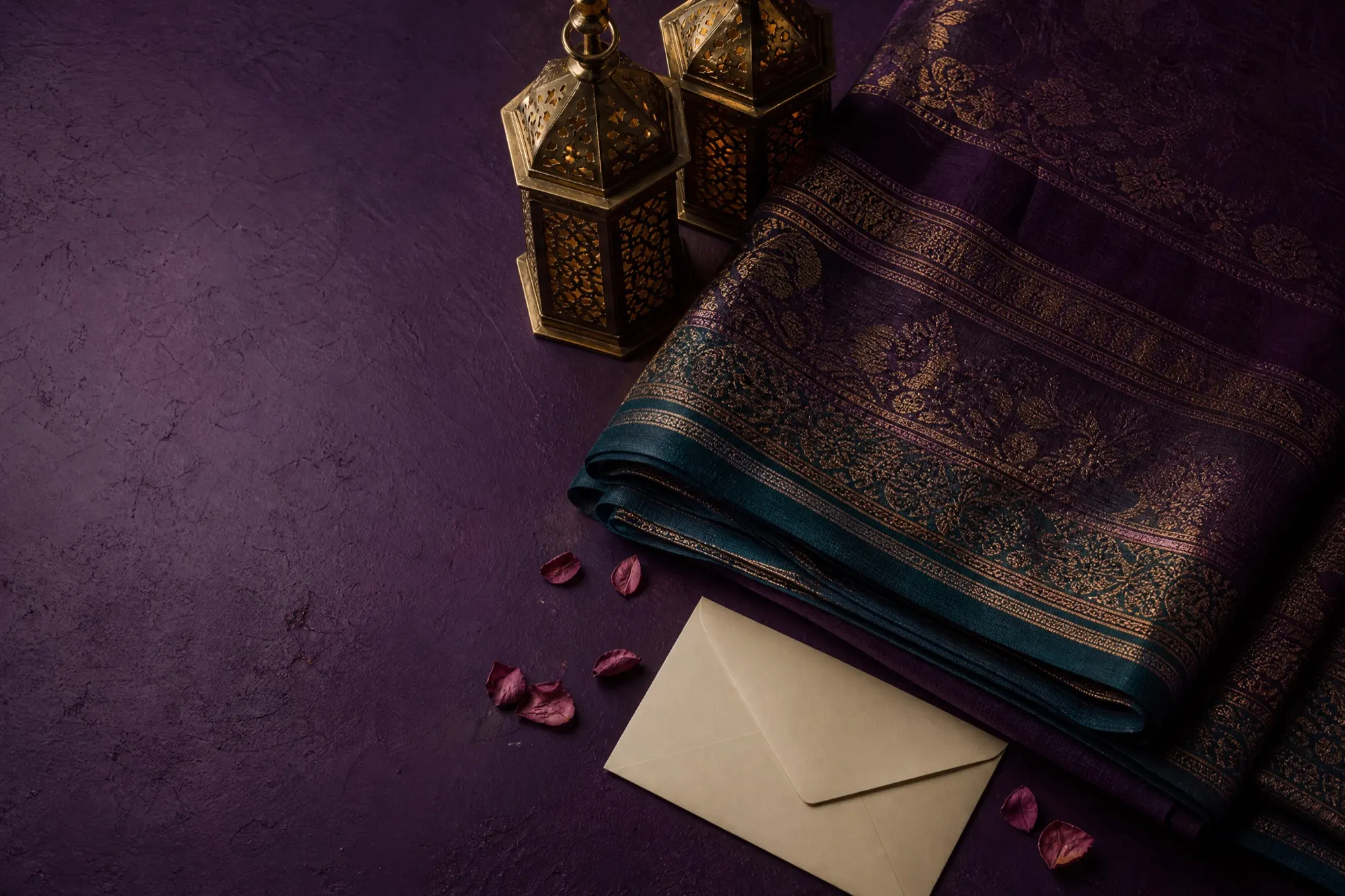 Folded brocade in plum and teal rests beside brass lanterns and a sealed envelope.